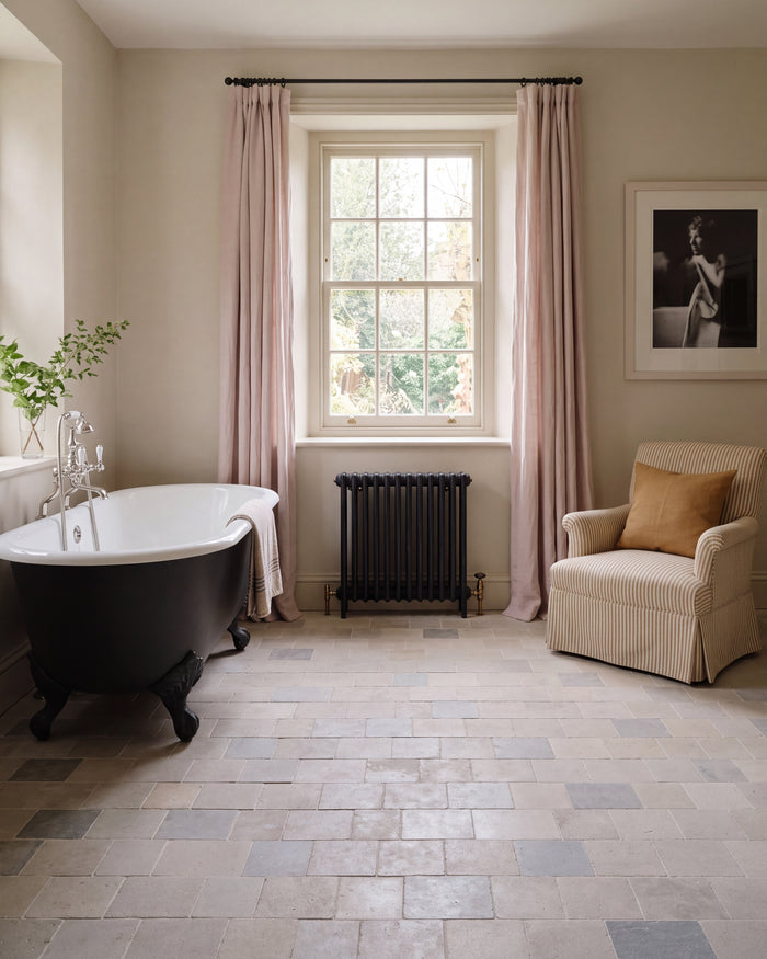 Castilan Limestone Cobbles Tumbled laid in a running bond across a bathroom floor with a black roll-top bath, cast iron radiator and ticking stripe armchair